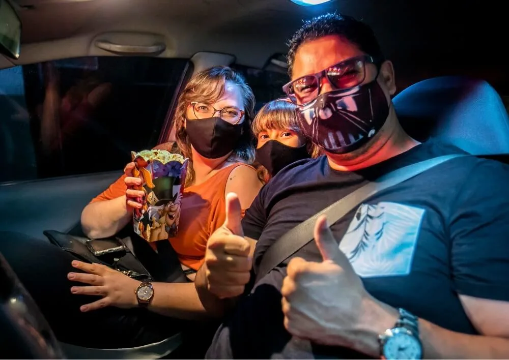 Família de máscara com pipoca, fazendo sinal de positivo dentro do carro